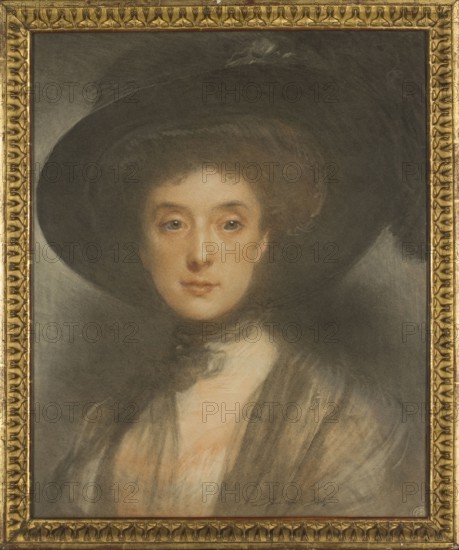 Portrait of a Woman in a Black Hat; Ludomir Janowski 1862–1939, painter; female portrait, ca. 1910; drawing, pastel; height 59 cm, width 47.8 cm; costume dated 1900–1915; hats headgear; Poland culture, A framed portrait of a woman wearing a large dark hat and a dark shawl, her face and neck shown with warm skin tones and subtle blush, light brown hair peeking from beneath the hat, soft blue-gray eyes, and a gentle neutral expression; the background and much of the hat and clothing are in deep gray and black shades with some muted brown and beige highlights, and the frame is an ornate gold color.