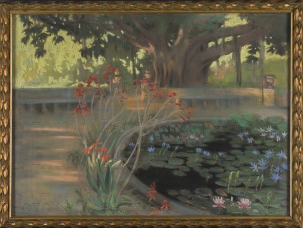 Lotuses in the Park in Palermo, Kazimierz Stabrowski 1869–1929, painter; pastel on paper, 1924; 30.0 × 39.5 cm. Depicts lotus flowers and pond in the botanical gardens Orto botanico of Palermo, Sicily, Italy; water landscape, gardens. Polish artist; Polish pastel., A framed painting showing a garden scene with a large tree in the background, dark brown and muted green trunk and branches, and a canopy of dark green and yellowish leaves. In front of the tree is a low stone wall in gray and beige tones that curves around a pond filled with dark water and many green lily pads. Several water lilies and small flowers in pale blue, light pink, and white float among the pads. Near the pond edge there are clusters of tall, slender plants with green leaves and red and orange blossoms. The ground and surrounding paths show muted shades of gray, brown, and soft peach, while distant foliage includes tones of light yellow-green and olive. The painting is set within an ornate gold frame.