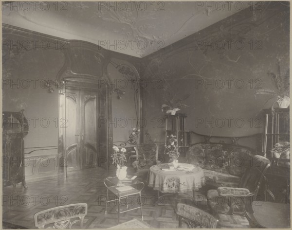 Lódz. Maurycy Poznanski's Palace. View of the drawing room with Art Nouveau decoration and furnishings, Piotrowski photographic studio; 1893–1934 author. Architecture; interiors; palaces; furniture; fabrics; paneling; stucco; doors; Art Nouveau style; modernism; Poland; c. 1900. Photography; photographic print: height 23.5 cm, width 29.6 cm; backing: height 35.1 cm, width 40.6 cm; print glued across the entire surface to decorative cardboard., The image shows an interior room with wooden paneled walls and carved wooden double doors, ornate molded ceiling details, a patterned wooden floor, a curved upholstered sofa and matching armchairs, a round table covered with a decorative cloth and a few items on top, a small metal-framed side table with a potted plant, several potted plants on tall stands, a curved wooden display cabinet, wall-mounted light fixtures, and a few additional chairs visible at the edges; visible colors include shades of brown, beige, cream, and muted green.