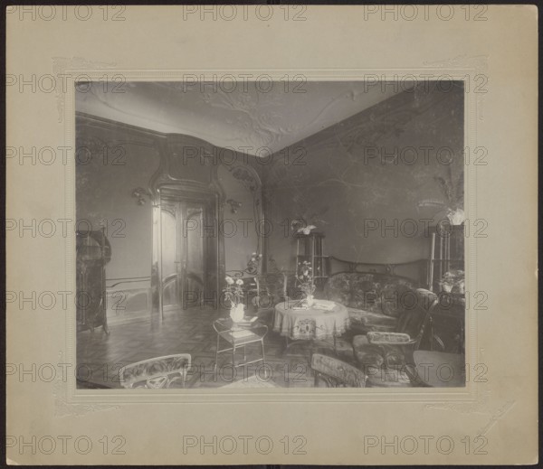 Lódz. Maurycy Poznanski's Palace . View of the drawing room with Art Nouveau decoration and furnishings, Piotrowski photographic studio; 1893–1934 author, architecture, interiors, Polish architecture, palaces, furniture, Lódz Lódz Voivodeship, fabrics, paneling, stucco, doors, Art Nouveau style, Poland culture, modernism style, print glued across the entire surface to decorative cardboard, c. 1900, Lódz place of origin, photography, photographic print: height 23.5 cm, width 29.6 cm; backing: height 35.1 cm, width 40.6 cm, The image shows an interior room with ornate woodwork and carved double doors, a decorative ceiling, patterned wooden floor, and curved walls; there is a group of upholstered chairs and a curved sofa arranged around a round table covered with a cloth, with vases of flowers on the table and on small side tables; several tall plant stands with potted plants are visible near the walls, and a small glass-topped table with a vase sits closer to the foreground; the visible ...
