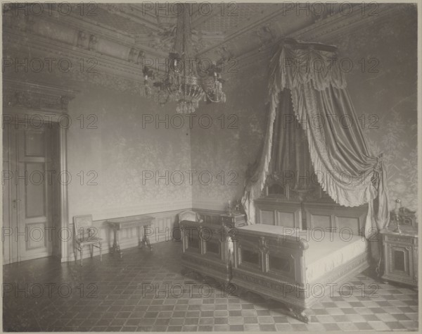 Lódz. Maurycy Poznanski's Palace . Bedroom interior with a double four-poster bed, Piotrowski photographic studio; 1893–1934 author, architecture, Lódz Lódz Voivodeship, Polish architecture, interiors, palaces, furniture, wallpapers, stuccoes, beds, bedrooms, chandeliers, Poland culture, modernism style, print glued across the entire surface to decorative cardboard, c. 1900, Lódz, nature photography, photographic print: height 23.2 cm, width 29.5 cm; backing: height 35.1 cm, width 40.6 cm, A large ornate bedroom with a high ceiling and decorative molding, shown in tones of sepia and muted brown. A carved wooden bed with an elaborate headboard and footboard sits to the right, draped with heavy curtains and a decorative canopy of flowing fabric in similar brown tones. A chandelier hangs from the ceiling above, with glass or crystal drops catching light. The patterned walls feature a floral motif, and the floor shows a checkerboard tile pattern. A wooden door is visible on the left, and n...