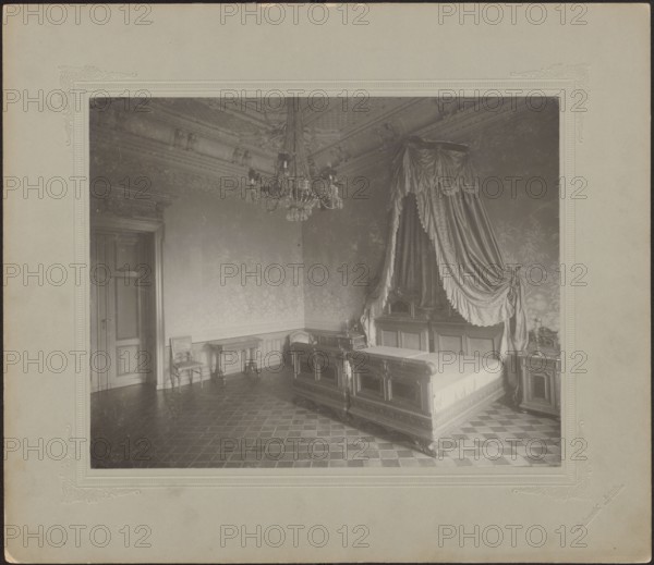 Lódz. Maurycy Poznanski's Palace . Bedroom interior with a double four-poster bed, Piotrowski photographic studio; 1893–1934 author, architecture, Lódz Lódz Voivodeship, Polish architecture, interiors, palaces, furniture, Pomian, Jan, wallpapers, stuccoes, beds, bedrooms, chandeliers, Poland culture, modernism style, print glued across the entire surface to decorative cardboard, c. 1900, Lódz, nature photography, photographic print: height 23.2 cm, width 29.5 cm; backing: height 35.1 cm, width 40.6 cm, The image shows an interior room with a large ornate bed draped with heavy fabric canopy and curtains, a decorative chandelier hanging from a detailed ceiling, patterned wallpaper, a tiled floor with a geometric pattern, a wooden door, a small table and chair against a wall, and a bedside table with objects on it; visible colors include shades of gray, black, white, and muted beige.