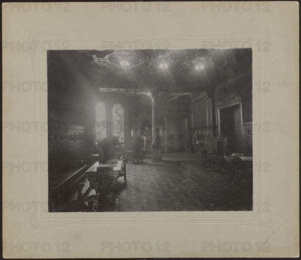 Lodz. Maurycy Poznanski Palace. Interior of a study in the Neo-Renaissance style, with a stained-glass window in the Art Nouveau style, interiors, architecture, Polish architecture, palaces, furniture, Lódz Lódz Voivodeship, panelling, stuccoes, coffers, studies, Neo-Renaissance style, Art Nouveau style, stained-glass windows, Poland culture, modernism style, print glued across the entire surface to decorative cardboard, ca. 1900, Lódz place of origin, photography, photography from nature, photographic print: height 22.2 cm, width 28 cm, backing: height 35.1 cm, width 40.6 cm, The image shows an interior room with ornate furniture and architectural details. A large central column rises from a patterned floor toward a decorative ceiling with visible light fixtures. Several high-backed carved chairs and tables with intricate legs are arranged around the room. Tall arched windows on one wall allow light to enter, casting highlights and shadows across the space. The walls feature molded pa...