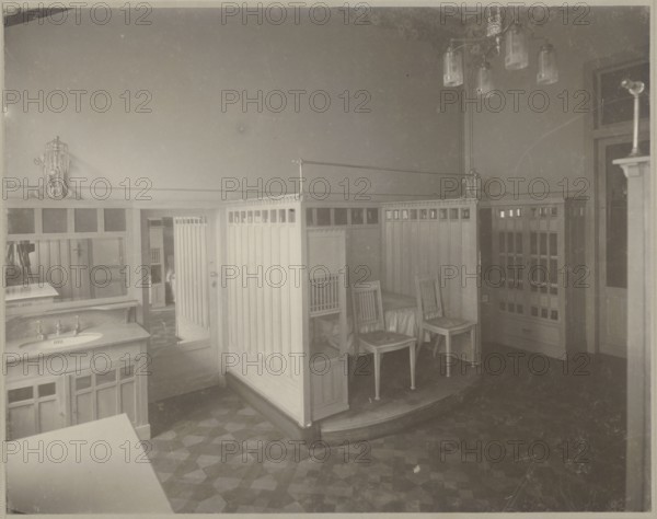 Lódz. Maurycy Poznanski's Palace. Interior of the bathing room. Photographic print by Piotrowski photographic studio; ca. 1893–1934. Art Nouveau / modernism style; Polish architecture; palace interior, bathroom. Print glued across the entire surface to decorative cardboard; photographic print: height 23.4 cm, width 29.8 cm; backing: height 35.1 cm, width 40.6 cm. ca. 1900. Lódz, Poland., The image shows an interior room with wooden panel partitions forming a group of small enclosed seating areas around a raised curved platform; a few wooden chairs with vertical slat backs are placed inside the enclosures and a small bed or cushioned seat is visible in one enclosure. A wooden sink cabinet with faucets and a mirror above it appears on the left, and additional built-in wooden cabinetry with glass panes is visible along the right wall. A multi-bulb hanging light fixture is suspended from the ceiling, and a decorative fixture or bracket is mounted on the left wall. The floor has a patterned...