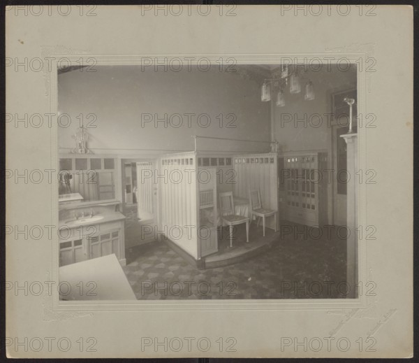 Lódz. Maurycy Poznanski's Palace . Interior of the bathing room. Piotrowski photographic studio; 1893–1934 author. Polish architecture, furniture, Art Nouveau, modernism; palaces, palace interiors, bathrooms; Lódz Lódz Voivodeship, Poland. Print glued across the entire surface to decorative cardboard, ca. 1900. Photography, photographic print: height 23.4 cm, width 29.8 cm; backing: height 35.1 cm, width 40.6 cm., The image shows an interior room with a central wooden partitioned structure forming a curved enclosure containing a few wooden chairs and a bed-like area; surrounding the enclosure are wooden cabinets and a countertop with a sink, all in light wood tones. The floor has a patterned surface in darker shades, and the walls are plain and light-colored. A hanging light fixture with several glass shades is visible near the ceiling, and there are doorways and tall cabinets along the walls. Overall colors visible include light beige and tan wood tones, darker brown and gray floor to...