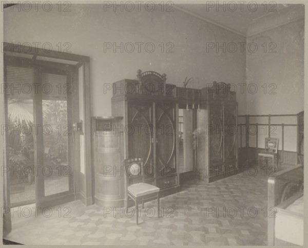 Lódz. Maurycy Poznanski's Palace . Interior of a bedroom, in the Art Nouveau style - view of the wall with wardrobes and exit to the orangery. Architecture, furniture, wardrobes, palaces, Art Nouveau, bedrooms, interiors, modernism; Pomian, Jan maker?. Photograph, print glued across the entire surface to decorative cardboard, c. 1900. Photography; photographic print: height 23.3 cm, width 29.2 cm; backing: height 35.1 cm, width 40.6 cm., The image shows an interior room with a large dark wooden wardrobe unit along one wall, featuring ornate top detailing and oval paneling, flanked by a cylindrical wooden cabinet. A wooden chair with a light-colored seat cushion sits in front of the wardrobe, and a second similar chair is placed near the corner. The floor has a patterned parquet design in shades of brown. To the left, tall glass doors open onto a space with visible large leafy plants in green. The walls are light-colored, and there is a dark wooden railing or wainscoting along the right...