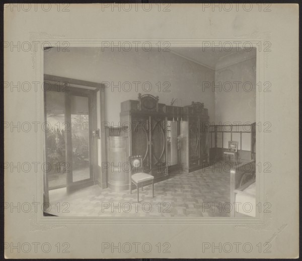 Lódz. Maurycy Poznanski's Palace . Interior of a bedroom, in the Art Nouveau style—view of the wall with wardrobes and exit to the orangery. Piotrowski photographic studio; 1893–1934 author. Architecture; furniture; wardrobes. Art Nouveau; modernism. Bedrooms; interiors. Poland; Lódz place of origin. Photography, nature photography. Photographic print print glued across the entire surface to decorative cardboard, c. 1900. Photographic print: height 23.3 cm, width 29.2 cm; backing: height 35.1 cm, width 40.6 cm., A room with pale walls and a patterned floor in shades of brown and tan containing a group of dark wooden cabinets with decorative tops and mirrored panels, a few wooden chairs with light upholstery, and a cylindrical wooden cabinet; a set of glass doors with dark frames opens to an area with leafy plants visible outside, and a low wooden railing runs along one wall; overall colors visible include brown, tan, beige, black, and pale grey.