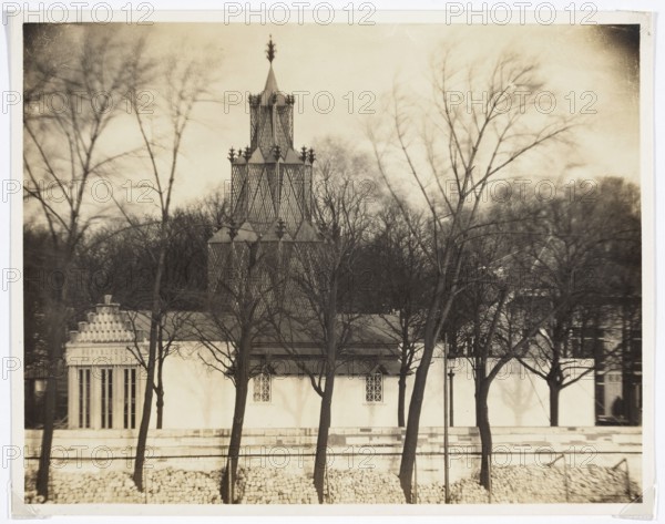 International Exhibition of Decorative Arts and Modern Industries Exposition Internationale des Arts Décoratifs et Industriels Modernes, Paris, 1925. Polish Pavilion designed by Józef Czajkowski — general view from the Seine. Photograph, 1925; photographer: REP Paris photographic studio. Subject: exhibitions; world exhibitions; Paris architecture; Polish architecture. Maker: Józef Czajkowski 1872–1947. Photography from nature. Loose print; height 14.5 cm, width 18.5 cm., A sepia-toned photograph shows a building with a tall, ornate, multi-tiered tower featuring lattice-like patterns, finial decorations, and pointed tops, set behind a row of leafless trees with thin, dark trunks and branches. In front of the building is a low wall and a rocky embankment. The building façade is light colored with a few small, diamond-patterned windows and a darker roofline. The background contains a dense grouping of bare trees and an overcast sky. Visible colors include shades of brown, beige, black, an...
