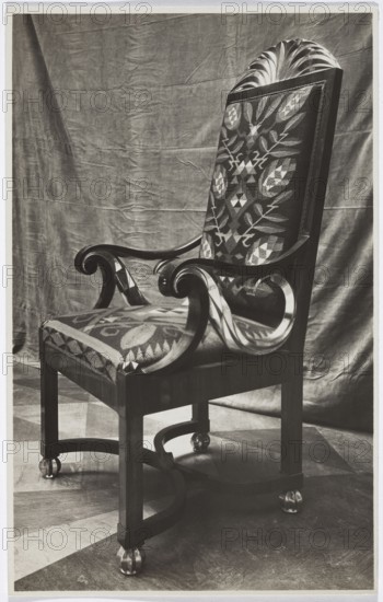 Armchair with kilim upholstery from the study designed by Józef Czajkowski, exhibited in the Polish Pavilion at the International Exhibition of Decorative Arts and Modern Industries Exposition Internationale des Arts Décoratifs et Industriels Modernes, Paris, 1925. Photograph, 1925; photographer: REP Paris photographic studio. Subject: furniture — armchair; materials: kilim upholstery; designer: Józef Czajkowski 1872–1947. Dimensions: 21.6 cm height, 13.6 cm width., A single ornate wooden chair is shown against a draped fabric backdrop. The chair has a tall, upholstered back with a geometric and floral patterned fabric and a cushioned seat covered in the same patterned material; the upholstery shows contrasting tones. The wooden arms curve into scroll shapes and have polished surfaces that catch light. The chair legs are straight and connected by curved stretchers that form a circular base element, and each leg rests on a rounded ornamental foot. The background fabric appears wrinkled ...