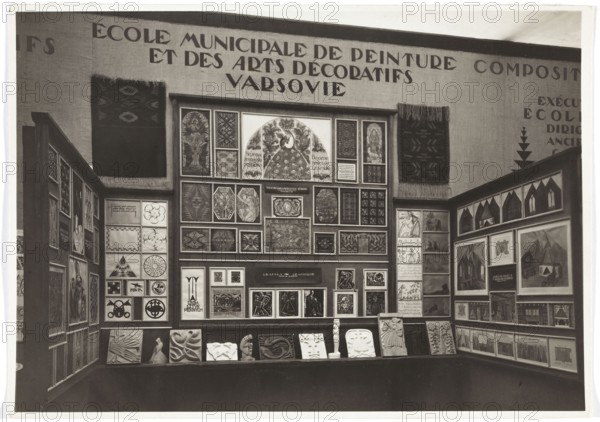 International Exhibition of Decorative Arts and Modern Industry Exposition Internationale des Arts Décoratifs et Industriels Modernes, Paris, 1925. Polish exposition in the Teaching Department of the Grand Palais — exhibition of the Municipal School of Decorative Arts and Painting in Warsaw. Photo REP Paris; photographic studio; fl. 1925. Exhibitions; world exhibitions; interiors; Paris France; Municipal School of Decorative Arts and Painting Warsaw; 1920–1939. Kilims; artistic fabrics; art schools; decorative arts; graphic art. Loose print, 1925; photography; nature photography. Height 16.0 cm, width 22.5 cm., A museum-like display of framed artworks arranged on three walls, with a central panel densely filled with a grid of varied rectangular and square pieces and two side panels showing framed images and decorative panels; a group of sculptural reliefs and small carved objects sit on a low shelf along the front. The images include stylized figurative scenes, geometric patterns, orna...