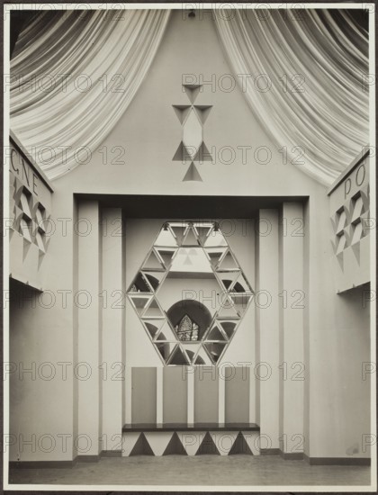 Vestibule designed by Wojciech Jastrzebowski for the Polish exhibition in the Interior Design Department at the International Exhibition of Decorative Arts and Modern Industries Exposition Internationale des Arts Décoratifs et Industriels Modernes, Paris, 1925. Photograph photographic studio REP, Paris, fl. 1925. Interiors; architecture; Polish architecture; mirrors. Photograph: print glued at top edge of album card, loose card; photographic print: 22.2 × 17 cm; cardboard: 33.1 × 40.4 cm., A symmetrical interior scene showing a central geometric installation with a large hexagonal frame made of triangular mirror panels, positioned within a recessed rectangular alcove flanked by vertical pilasters; above the alcove a triangular motif of raised triangular shapes is visible on a plain wall, and draped fabric sweeps down from the top corners creating curved folds. The foreground shows a bench-like ledge with alternating light and dark triangular patterns beneath it, and the walls on either...