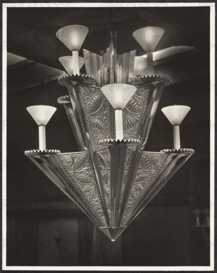 Chandelier in the workroom, designed by Mieczyslaw Kotarbinski, at the International Exhibition of Decorative Arts and Modern Industries Exposition Internationale des Arts Décoratifs et Industriels Modernes, Paris, 1925. Photograph of exhibition interior; photographic print, height 17.6 cm, width 13.9 cm; backing height 33.1 cm, width 40.4 cm., A decorative chandelier fills the frame, showing a layered, tiered metal structure with ornate embossed patterns and scalloped edges, topped by several frosted, cone-shaped glass shades mounted on slender cylindrical stems. The metalwork displays intricate, repeating leaf and geometric motifs with raised ridges and polished reflective surfaces. The glass shades are smooth and opaque, appearing lighter against the darker background. The scene contains tones of black, white, and various shades of gray, with highlights on the metal and glass and deep shadows behind the fixture.