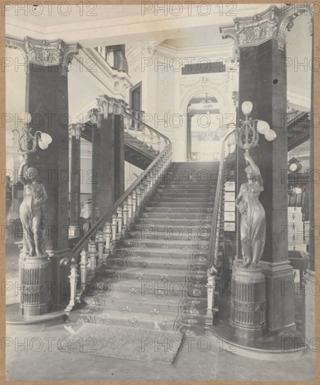 Staircase in the building of the Boguslaw Herse Fashion House at 150 Marszalkowska Street in Warsaw, Boguslaw Herse Fashion House Warsaw; 1868–1936, fashion, interiors, shops, fashion houses, Warsaw Masovian Voivodeship, tenement houses, Art Nouveau, Marszalkowska street, Poland, modernism, print glued across the entire surface onto a museum backing, ca. 1910, photograph from nature, photograph, height 26.7 cm, width 21.8 cm, A grand interior scene showing a wide central staircase with a patterned runner carpet and carved wooden handrails flanked by tall ornate columns. A pair of sculpted female statues stand on decorative pedestals at the base of the staircase, each holding multi-globe light fixtures. The staircase rises to a landing with balustrades and a large arched doorway visible above, with decorative molding and relief work on the walls. There is a rectangular mat at the foot of the stairs on a smooth floor. Visible colors include shades of gray, black, and off-white, with vari...