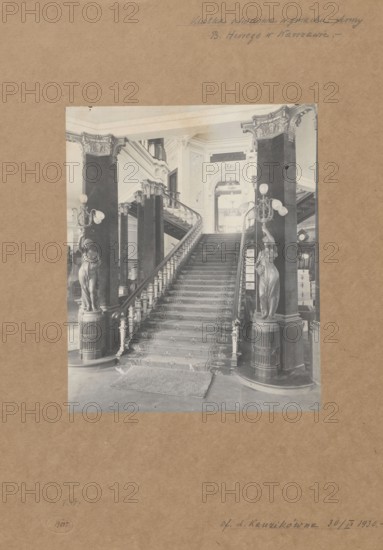 Staircase in the building of the Boguslaw Herse Fashion House at 150 Marszalkowska Street in Warsaw, unknown photographer/creator, Boguslaw Herse Fashion House Warsaw; 1868–1936, fashion, interiors, shops, fashion houses, Warsaw Masovian Voivodeship, tenement houses, Art Nouveau and modernism styles, Marszalkowska street, Poland, ca. 1910, photograph from nature, print glued across the entire surface onto a museum backing, photograph, height 26.7 cm, width 21.8 cm, A large sepia-toned photograph mounted on brown cardstock shows an ornate interior stairway flanked by two tall dark columns topped with decorative capitals and sculpted female figure lamp bases holding multi-globe lamps; the central staircase is carpeted with a patterned runner and has carved balustrades and newel posts, leading up to an arched opening with decorative moldings and relief work; a small rectangular rug lies at the base of the stairs on a smooth floor, and additional architectural details such as railings, col...