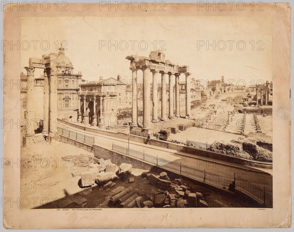 Roma. Foro Romano preso dal Campidoglio, James Anderson 1813–1877, photographer; Anderson Rome, publisher. Subjects: Forum Romanum Rome; architecture—ancient, Italian, foreign; Rome, Italy. Medium: photographic print glued across the entire surface onto thick cardboard backing. Date: ca. 1880. Dimensions: photographic print: 42.1 cm × 55.0 cm; cardboard: 48.9 cm × 62.5 cm., The image shows an open archaeological site with ruined classical columns and stone fragments, with a row of tall standing columns in the center and several shorter columns and ruins scattered around. A road or pathway with metal railings runs through the middle foreground, curving slightly and casting shadows. Buildings with ornate facades and a domed structure appear behind the ruins on the left, and more ruins and distant buildings extend into the background on the right. The ground and stone surfaces are shades of beige and tan, the columns and buildings are light beige, the railings and shadows are darker brown...