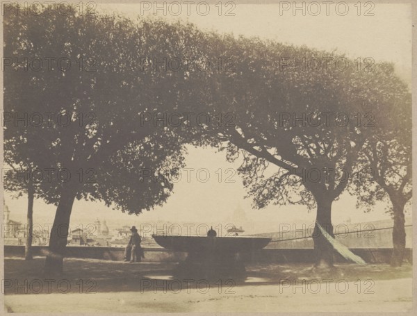 Fountain in the gardens of the French Academy at Villa Medici on Pincio Hill, Rome. View towards St. Peter's Basilica. Photographer: Giacomo Caneva 1813–1865. Subjects: architecture, fountains, city panorama, Villa Medici, Monte Pincio, Vatican, St. Peter's Basilica. Ca. 1852. Photography from nature; photographic print glued to cardboard. Print: 20.0 × 26.6 cm; cardboard: 24.6 × 34.5 cm., A sepia-toned scene shows a shaded park area with a group of large trees forming a canopy across the upper part of the image, their dark foliage contrasting with a pale sky. Under the trees, silhouettes of tree trunks and branches create dense shadowed shapes. In the middle ground there is a wide, shallow fountain basin with a rounded silhouette, and near it a person wearing a hat standing by the low stone balustrade, looking toward a distant cityscape that appears faint and light against the horizon. The foreground is an open, sunlit ground area in muted beige tones, and a lighter path or strip runs...