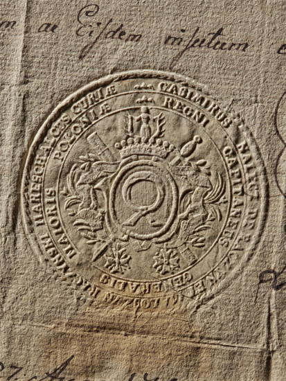 Seal of Kazimierz Raczynski, Starost General of Greater Poland. Imprint of the seal. In an oval decorative cartouche, the Nalecz coat of arms with a jewel above a nine-baton crown. Behind the cartouche, a crossed karabela and a scabbard; below, on ribbons, two orders. In the double rim an inscription: CASIMIRUS NALECZ DE RACZYN ET MALOSZYN RACZYNSKIMARESCHALCUS CURIAE //REGNI CAPITANEUS GENERALIS MAIORIS POLONIAE, 1785. Poland., A close-up of an embossed circular seal pressed into textured paper, showing raised ornamental detailing including a central circular emblem with surrounding decorative flourishes, a crown-like motif near the top, and floral or star-shaped decorations near the lower area; the paper has a rough, fibrous surface with visible creases and folds, and there is cursive handwriting partially visible near the top and edges. The visible colors are beige, tan, brown, and darker brown in the inked handwriting.