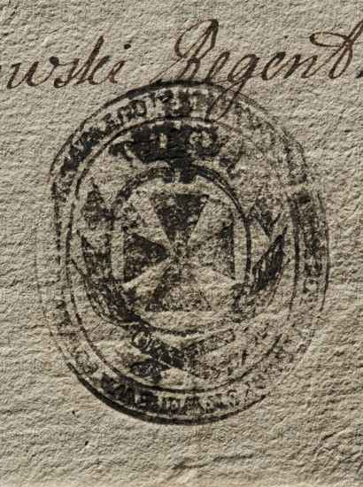 Seal of the Civil-Military Order Commission of the Braclaw Voivodeship and County; oval ink impression of the seal. On an oval crowned shield, a knight's cross—the coat of arms of the Braclaw Voivodeship—surrounded by a panoply. Illegible inscription on the rim. 1790, Poland; seals attached to documents; documents; official seals; seals with provincial coats of arms; maker: Zurakowski, Witold (?-1927)., A textured beige paper surface with a faded circular dark stamp that contains a central shield-like shape and a cross-like motif, surrounded by wreath-like decorative elements and an outer ring; above the stamp is a sweep of cursive handwriting in brown ink. Colors visible include beige, dark brown, and black.