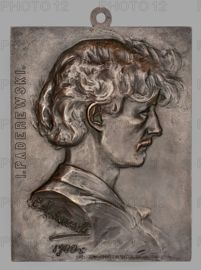 Czeslaw Makowski (1873–1921), bust of Ignacy Jan Paderewski (1860–1941), male bust in right profile with slightly wavy, longer hair falling on the forehead and a moustache; inscription along the left edge: 'I. PADEREWSKI.', dated 1900. Plaque, Poland, Warsaw. Dimensions: 1205 mm and 265 mm., A rectangular metal plaque with a loop at the top shows a raised profile of a young person facing right with wavy hair and a faint mustache, the hair rendered with textured flowing locks in brownish bronze and darker patina, with hints of green oxidation near the ear; the figure wears a collared garment with sculpted folds in muted bronze and gray tones, and the background of the plaque is a slightly mottled gray-brown metal with areas of darker and lighter shading.