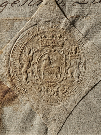 Seal of Count Ignacy Zaluski, Starost of Checiny. Round dry impression of the seal. Crowned cartouche with the Junosza coat of arms supported by two lions. In the rim is an illegible inscription. 1775, Poland., A close-up of textured paper showing an embossed circular seal with ornate heraldic elements, including a central shield flanked by two standing figures and topped with a crown, all rendered in raised relief; the paper shows creases and layered edges, with visible wear and uneven surface fibers. The colors visible are various shades of beige, tan, brown, and a few darker brown accents.