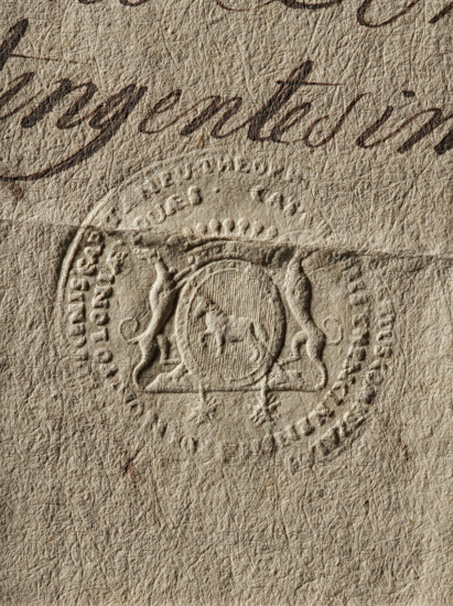 Seal of Teofil Count Zaluski, Starost of Checiny. Round seal impression of the Crown Court Treasurer Teofil Count Zaluski, Starost of Checiny. Crowned oval cartouche with the Junosza coat of arms supported by two hounds. Inscription on the double rim, largely illegible. 1789, Poland., A close-up of textured beige paper showing an embossed circular seal with a raised crest featuring animal figures and decorative flourishes, a central oval containing a small embossed animal, and surrounding embossed shapes; a segment of dark brown cursive ink appears near the top, and the image overall shows variations of beige, tan, and brown tones with visible fine fibrous texture and subtle shadows around the embossing.