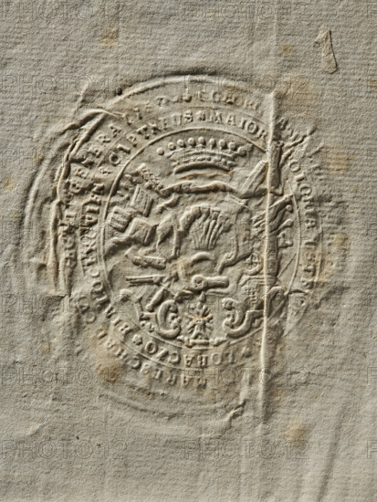 Seal of Jerzy August Wandalin Mniszech, starost general of Greater Poland; starost of Sanok, Bialocerkiew, Lubaczów. Oval impression of the seal. The coat of arms is placed in a rich cartouche under a crown, with a panoply behind the shield. The inscription on the double rim is partially illegible. 1778, Poland. Seal. Dimensions: 59.5 mm and 65.9 mm., The image shows an embossed circular seal pressed into textured paper, creating raised and recessed areas; the paper is mostly beige with variations of light tan and small darker brown spots, and the seal impression forms concentric rings with ornate decorative elements and sculpted shapes including a crown-like motif and stylized figures and flourishes; the surface has creases and subtle shadows that emphasize the relief, and the overall appearance is aged and weathered with a few darker stains and slight discolorations.