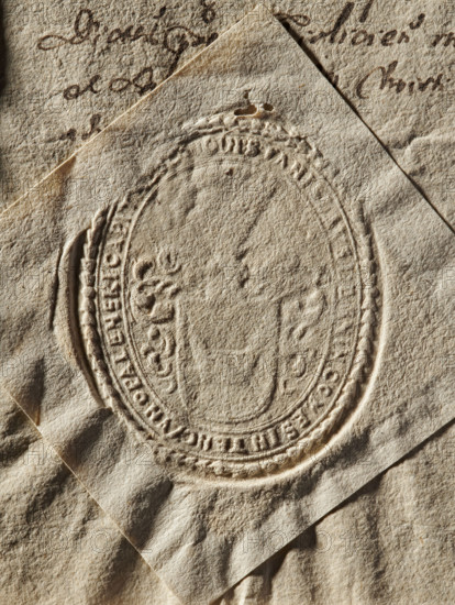 Seal of Stanislaw Opalinski, starost of Korczyna (1647–1704). Oval impression of a private/document seal showing the Lodzia coat of arms on a shield with a crowned helmet with a jewel and labrums. Partially illegible rim inscription: 'STANISLAVS DE , COMES IN TENCZYN OPALENERI CAPITANEUS, 1676.' Poland; seal. Dimensions: 44.2 mm and 51.8 mm., The image shows an embossed oval seal impressed into textured, aged paper with a rough, fibrous surface in beige and tan tones, layered over another sheet with cursive ink markings partly visible; the seal creates raised and recessed areas forming decorative borders and internal shapes, with shadows and highlights emphasizing creases and folds in the paper, and the overall color palette includes shades of beige, tan, brown, and off-white.