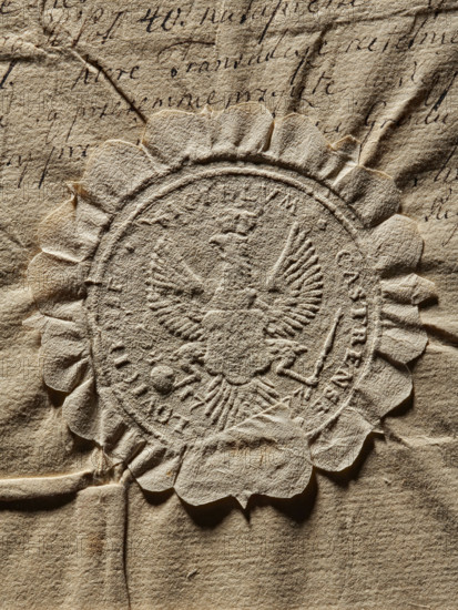 Seal of the starost of the town of Lowicz (ca. 1777). Round imprint showing a crowned White Eagle holding a scepter and an orb; on its chest a shield, probably with the coat of arms of Lowicz. Rim inscription: SIGILLVM CASTRENSE LOVICIENSE, 1777. Seal (Poland)., The image shows a close-up of a textured, crumpled beige paper surface with an embossed circular seal featuring a raised design of an eagle with outstretched wings and decorative scalloped edges; the paper displays creases, folds, and uneven fibers in shades of tan, light brown, and cream.