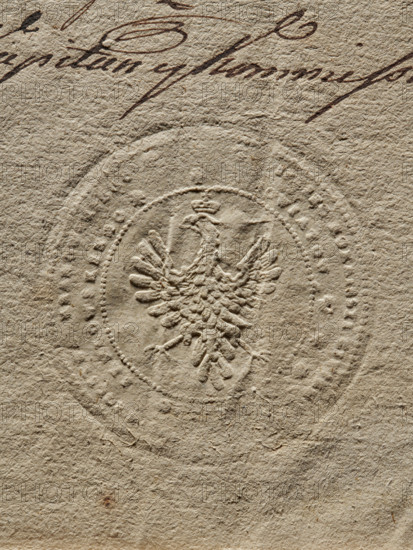 Civil-Military Order Commission of the Kraków Voivodeship, Lelów County and the Duchy of Kraków; Civil-Military Order Commission; Zurakowski, Witold (?-1927) — documents, official seals, seals with an eagle; Round seal of the Civil-Military Order Commission of the Kraków Voivodeship, Lelów County and the Duchy of Kraków. Polish eagle with a crown. In the rim the inscription: 'PIECZEC KOMMISSYI CIWILNO MILITOWEY WOJEWODZTWARAKOWSKIEGO // POWIATOW XIESKIEGO I LELOWSKIEGO', 1790, Poland, seal, A close-up view of textured beige paper showing an embossed circular seal with a raised heraldic bird at its center, the bird featuring outstretched wings and a small crown. The paper surface displays a fibrous, rough texture with variations in tone, and a few darker brown ink pen strokes appear near the top edge. The visible colors are beige, tan, and shades of brown.