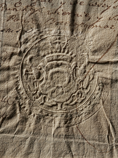 Starost of Pilzno seal, Starost of Pilzno (ca. 1767). Round seal impression. In a decorative cartouche, the Nalecz coat of arms with a jewel above a nine-baton crown. Illegible inscription in the double rim. 1767, Poland., The image shows a close-up of aged, textured paper in beige and tan tones with darker brown ink marks visible across the surface; the paper has several creases and folds and a raised, embossed circular seal with ornate, decorative patterns and scroll-like elements in the center, creating shadows and highlights that emphasize the rough fibrous texture.