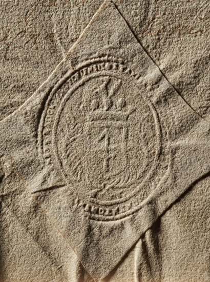Seal of Stanislaw Chometowski, Starost of Radom (1673–1728). Oval seal impression: a shield with the Lis coat of arms, a jewel above a rank crown, surrounded by two palm branches tied with a ribbon at the bottom. Inscription on the rim largely illegible. 1699, Poland. Seal; dimensions 42.5 mm and 50 mm., A textured sheet of paper in beige and tan tones lies with a diamond-shaped overlay that shows an embossed circular seal; the embossing creates raised and recessed patterns including a central emblem and surrounding decorative rings, and the paper surface has a rough fibrous texture with creases and shadows in shades of beige, tan, brown, and cream.