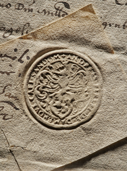 Round seal impression of Andrzej Firlej, Starost of Sandomierz. Shield with the Lewart coat of arms with very rich labres and a jewel above a crowned helmet. In the rim, an inscription: + ANDREAS FIRLEI DE DAMBROVI CAPI SANDOM +, 1577, Poland., A close-up of textured, off-white paper with a raised circular embossed seal in the center showing ornate, curving designs and a heraldic lion motif; the paper has a rough, fibrous surface with creases and overlapping torn edges. There are visible dark brown ink marks and scribbles on parts of the paper, and the overall colors include shades of beige, cream, brown, and tan.