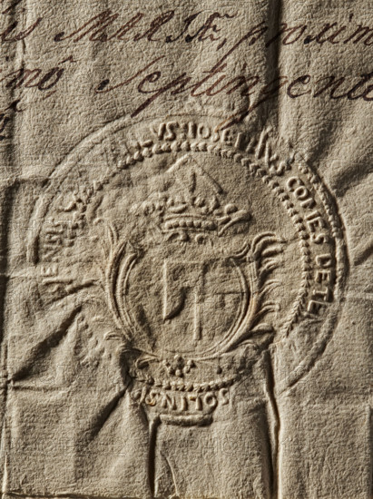 Seal of Józef Kanty Ossolinski, starost of Sandomierz (1707–1780). Round impression of a seal: in a round cartouche entwined with two palm branches is the Topór coat of arms with a jewel above a rank crown. In the rim is a partly illegible inscription: IOSEPHUS COMES DE TENCZYN OSSOLINSK, ENSIS CA, ANEVS., 1765, Poland. Function: stamp; private/document seal; starost's seal. Maker: Józef Kanty Ossolinski., The image shows a close-up of textured, aged paper with an embossed circular seal featuring a central shield-like emblem topped by a decorative crown and flanked by ornamental flourishes; the paper has visible creases and folds and a rough fibrous surface. There is handwritten ink near the top in a dark brown color, and the paper itself is a warm beige to light tan with subtle darker brown shadows in the embossed and creased areas.