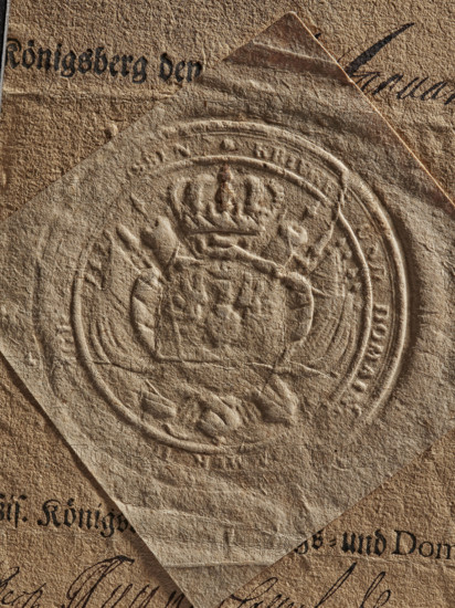 dry impression of an oval royal seal depicting an eagle with spread wings, royal camera seal, royal camera, documents, official seals, dry impression, document seals, seals with coats of arms, 1749, Poland, Królewiec, seal, A close-up of overlapping textured papers in shades of brown and tan, with a central embossed circular seal showing raised ornamental shapes including a crown-like form and decorative borders; the top paper is lighter tan and folded at one corner, revealing a darker brown sheet beneath with visible cursive ink strokes in black, and several areas of creasing, grainy fibers, and small dark specks across the surfaces.