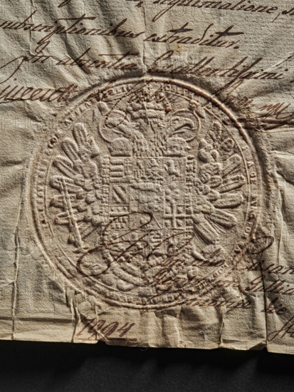 Great state seal; impression of the great state seal; official document seal; multi-field coat of arms with talons, wings, and two eagle heads, between them a crown; 1789; Austria; maker: Witold Zurakowski (?-1927), A close-up of a textured, aged paper with an embossed circular seal featuring a complex heraldic design and an ornate border; the paper shows creases, folds, and slight tears along the bottom edge. Over the embossed area and surrounding paper there is flowing cursive handwriting applied with a dark brown ink. Visible colors include beige and tan tones of the paper, dark brown of the handwriting, and subtle darker shadows around the creases.