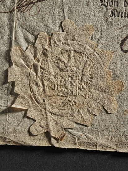 Dry impression of an official seal; coat of arms with wings and two eagle heads with crowns, 1802, Austria; official seal, document seals; Zurakowski, Witold (?-1927), A close-up view of an aged, textured paper seal affixed to an old sheet of paper, showing a scalloped, star-like edge and an embossed central design with raised and indented areas; the papers display a coarse, fibrous surface with creases and small tears. The visible colors are beige, tan, off-white, brown, and a dark gray-black background at the bottom.