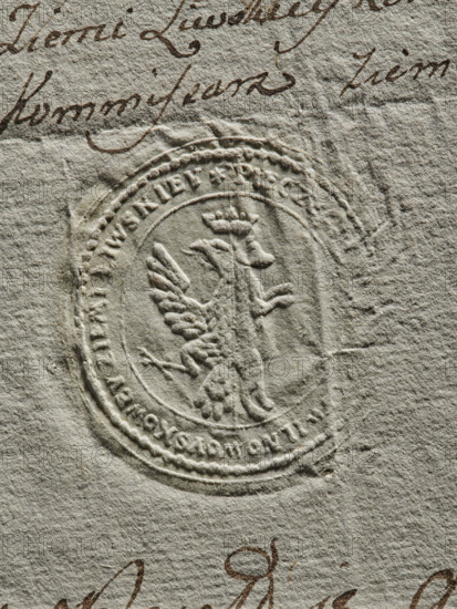Seal of the Civil-Military Order Commission of the Liw Land, dry impression, document seal. Round seal of the Civil-Military Order Commission of the Liw Land. In the field of the seal, a half-eagle and a south-bear under a common crown. Around the rim, the inscription: PIECZEC KOMMISSYI CYWILNO WOYSKOWEY ZIEM LIWSKIEY, 1790. Poland, Liw Land., A close-up of textured off-white paper with an embossed round seal featuring a raised winged creature wearing a crown at its center, surrounded by concentric circular ridges and decorative edging; there is brown cursive handwriting visible near the top and bottom edges, and the image shows subtle shadows and creases in the paper with visible fibers and a few darker smudges.