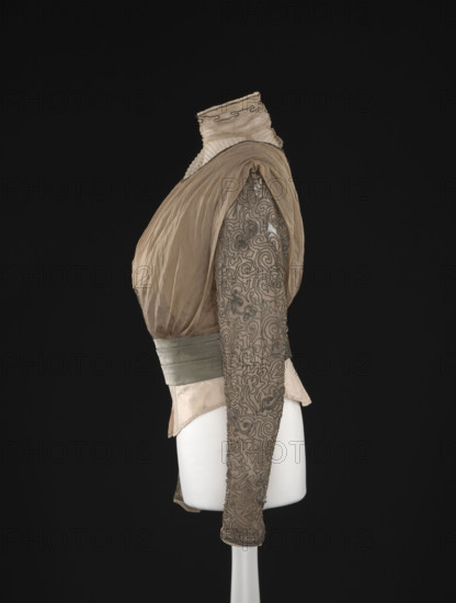 Blouse. Made of beige-gold muslin, short, bloused, fitted at the waist with a belt draped in three pleats of green-olive fabric, lined. At the front from the top, an insert of tulle sewn in small horizontal pleats, passing into a stand-up collar lined with muslin, decorated with a single sewn-on silver metal string. The remaining part is made of gathered, double-folded muslin with a semi-circular neckline finished with a similar string. The lower part is decorated with a rectangular element embroidered with a metal string in geometric motifs. The shoulders are gathered with bias-stitched, loosely fitted, gathered at the waist 'overlays' in a kind of cape. The back is similar to the front with a full closure. The long sleeves are slightly gathered at the top, narrow, and decorated with a sewn-on metal string in geometric motifs. The blouse has a fitted, sewn-in lining of light cream taffeta, stiffened with nine bonings. The blouse has a muslin bottom with a fragmentarily preserved gray ...