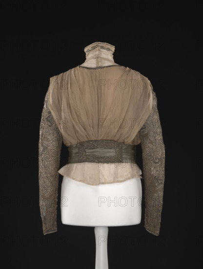 Blouse. Made of beige-gold muslin, short, bloused, fitted at the waist with a belt draped in three pleats of green-olive fabric, lined. At the front from the top, an insert of tulle sewn in small horizontal pleats, passing into a stand-up collar lined with muslin, decorated with a single sewn-on silver metal string. The remaining part is made of gathered, double-folded muslin with a semi-circular neckline finished with a similar string. The lower part is decorated with a rectangular element embroidered with a metal string in geometric motifs. The shoulders are gathered with bias-stitched, loosely fitted, gathered at the waist 'overlays' in a kind of cape. The back is similar to the front with a full closure. The long sleeves are slightly gathered at the top, narrow, and decorated with a sewn-on metal string in geometric motifs. The blouse has a fitted, sewn-in lining of light cream taffeta, stiffened with nine bonings. The blouse has a muslin bottom with a fragmentarily preserved gray ...
