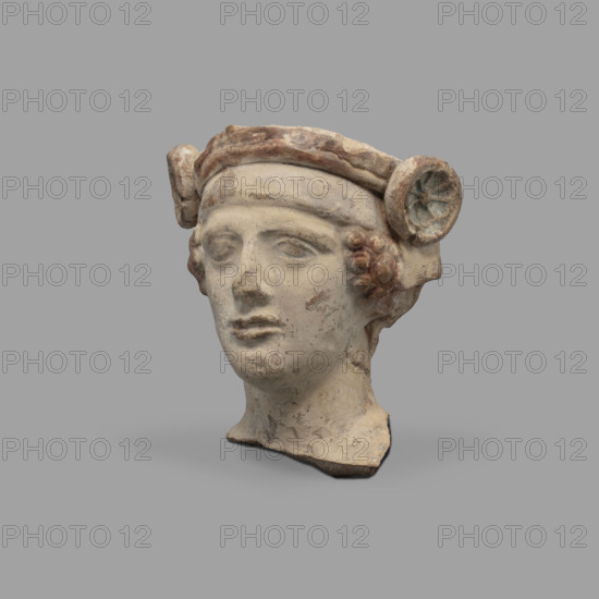 Head of a figurine of Dionysus. Head of a young man with a face devoid of [expression?], with a wide, compact mouth, a triangularly outlined face with carved cheeks, large eyes, a prominent nose and gently outlined eyebrows. On his head the youth wears a diadem decorated with rosettes on the sides and a thick wreath. Traces of red paint (cinnabar) on the hair, lips and diadem; blue on the rosettes (in the recesses). 5th–4th century BC. Taranto, Southern Italy (Italy). Terracotta figurine; height 8.7 cm, width 8.3 cm, depth 6.3 cm, weight 0.28 kg.