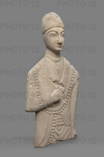 Fragment of a votive figure of a man. The fragment depicts a standing man dressed in a tutulus and himation. The outer edge of the himation is framed by an ornament of semicircular serrated edges. The torso is flattened; the front is carefully detailed while the back is flat. The right arm, bent at the elbow, rests on the chest; the right hand is lowered close to the torso, clenched into a fist. The face is oval and elongated with a small chin, thin lips, a long aquiline nose, slanted prominent eyes, arched eyebrows, and a low forehead. Conical headdress. Proto-Cypriot Style II, 600–480 BC (previous dating: 1st half of the 6th century BC). Sculpture, Cyprus. Height 28 cm, width 14 cm.