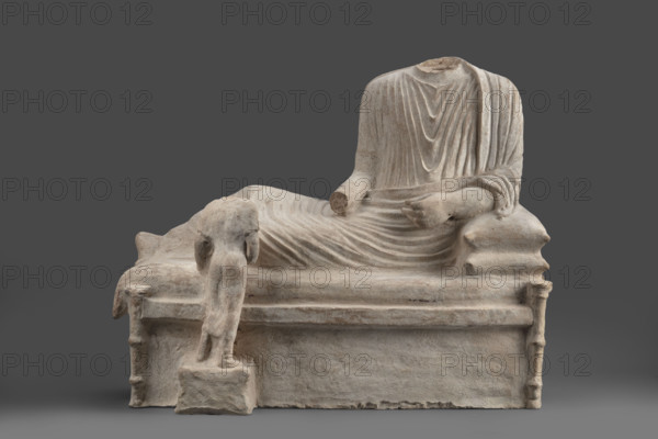 Depiction of a deceased person on a keline and a servant. The sculpture depicts a man banqueting in a semi-reclining position on a keline (bed). His upright torso is supported by his left elbow placed on two pillows. The man is dressed in a long, finely pleated chiton and himation. In front of the bed, at the side of the banqueter's legs, stands a small boy dressed in a chiton on a cuboid base. 4th century BC (dating before the 2019 revision: 450–375 BC; Cypriot-Classical Period). Marion. Sculpture, height 45 cm, width 53 cm, depth 24 cm, weight 13.03 kg.
