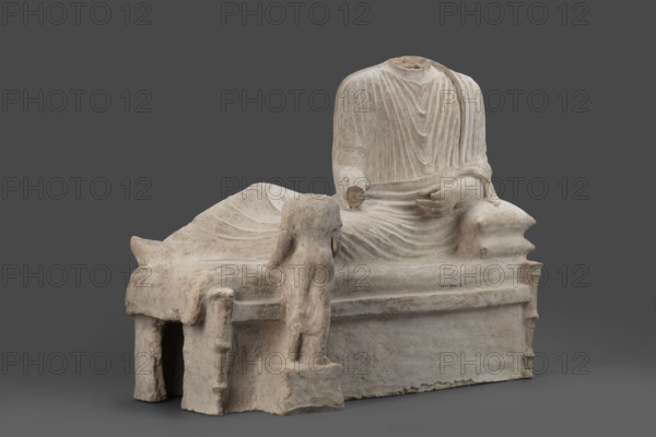 Depiction of a deceased person on a keline and a servant; banquet scene, Cypriot sculpture. The sculpture depicts a man banqueting in a semi-reclining position on a keline (bed). His upright torso is supported by his left elbow placed on two pillows. The man is dressed in a long, finely pleated chiton and himation. In front of the bed, at the side of the banqueter's legs, stands a small boy dressed in a chiton on a cuboid base. 4th century BC (dating before the 2019 revision: 450–375 BC; Cypriot-Classical Period). Marion. Sculpture. Height 45 cm; width 53 cm; depth 24 cm; weight 13.03 kg.