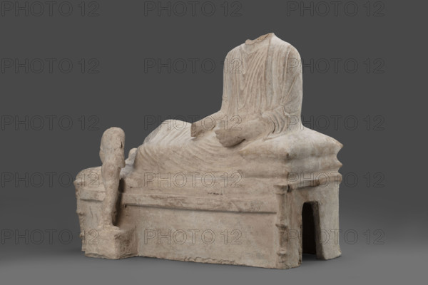 Depiction of a deceased person on a keline and a servant; banquet scene, Cypriot sculpture. The sculpture depicts a man banqueting in a semi-reclining position on a keline (bed). His upright torso is supported by his left elbow placed on two pillows. The man is dressed in a long, finely pleated chiton and himation. In front of the bed, at the side of the banqueter's legs, stands a small boy dressed in a chiton on a cuboid base. 4th century BC (dating before the 2019 revision: 450–375 BC; Cypriot-Classical Period). Marion. Sculpture; height 45 cm, width 53 cm, depth 24 cm, weight 13.03 kg.