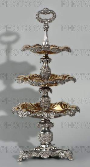 Etazera by Józef Fraget (Warsaw; silver and plated goods factory; 1824–1945). Three-tiered platter on a three-sided base with three knobbed plates; plated with gold on the outside and silver on the bottom, with silver-plated tin overlays on the edges. Finished with an oval decorative handle. Composed of two parts: the base and a plate holder inserted into a cylindrical opening. Marked with an oval Fraget mark dated 1843. Vessels, 1843, Poland, Warsaw. Dimensions: height 57 cm, width 25.9 cm.