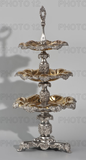 Etazera, Fraget, Józef (Warsaw; silver and plated goods factory; 1824–1945), tableware elements. Three-tiered platter on a three-sided base with three knobbed plates, plated with gold on the outside and silver on the bottom, with silver-plated tin overlays on the edges. Finished with an oval decorative handle. Composed of two parts: the base and a plate holder inserted into a cylindrical opening. Marked with an oval Fraget mark with the date 1843. Vessels, 1843, Poland, Warsaw. Height 57 cm; width 25.9 cm.