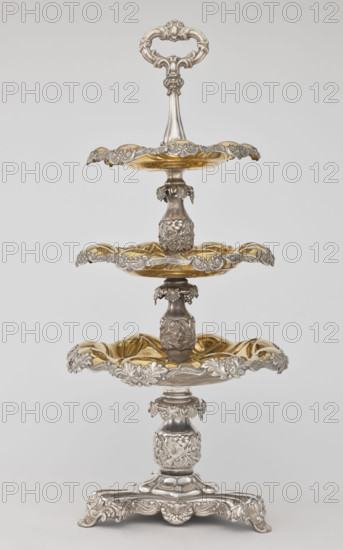 Etazera, Fraget, Józef (Warsaw; silver and plated goods factory; 1824–1945). Three-tiered platter on a three-sided base with three knobbed plates, gold-plated on the outside and silver on the underside, with silver-plated tin overlays on the edges. Finished with an oval decorative handle. Composed of two parts: the base and a plate holder inserted into a cylindrical opening. Marked with an oval Fraget mark dated 1843. Vessels, 1843, Poland, Warsaw. Dimensions: height 57 cm, width 25.9 cm.