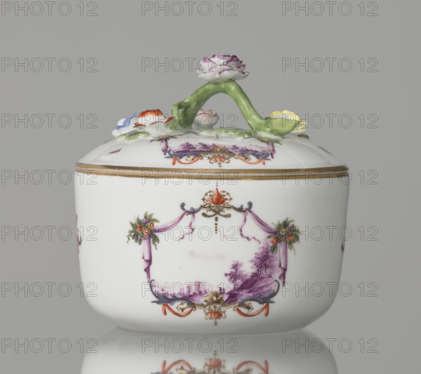 Sugar bowl with a lid and a branch handle, Koenigliche Porzellain Fabrique, Meissen (1710–1763), manufacturer. Cylindrical sugar bowl, rounded at the base, with a slightly arched lid and a plastic, colorfully painted branch handle with four flowers and leaves at the end of the side shoots: three forget-me-nots, two brick-red flowers and one yellow. On the sides of the body two cartouche frames and on the cover one, each framed by two purple draperies suspended from the sides on small flower bouquets and dry rocailles, topped at the top and bottom on the axis of symmetry by brick-red draperies suspended on plant branches. Within the cartouches are landscape miniatures with architecture and staffage painted in purple camaieu: 1) a fragment of a hill with trees, two figures, and a building in a valley; 2) two buildings among bushes; cover: 3) a plateau with a house and a hill in the distance on the right. Between the cartouches on the body are two colorful ornamental-floral motifs; on the...