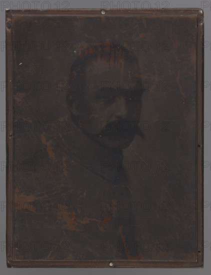 Portrait of Józef Pilsudski (1867–1935), 1924, graphic plate; height 12.8 cm, width 9.7 cm, thickness 2.1 cm; graphic matrix: height 12.7 cm, width 9.6 cm, thickness 0.2 cm; artist: Leon Wyczólkowski (1870–1927) — iconography., A dark, rectangular framed image showing a faded portrait of a person with short hair and a mustache, the face and upper body rendered in low contrast against a deep brown and black background; a few rusty reddish-brown marks and scratches are scattered across the surface, and the frame edges are visible in darker brown with small metallic fasteners at the top and bottom.