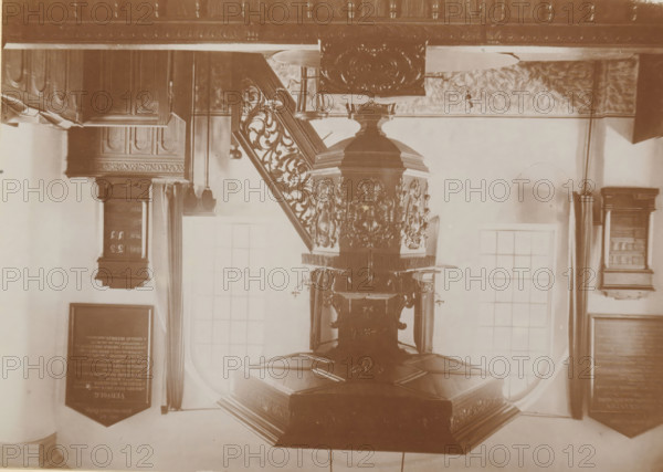 Pulpit in the Old St. Victor Church in Batenburg, photograph, page, photographer: Benjamin Charlé (possibly), Batenburg, c. 1903 - in or before 1908, paper, baryta paper, daylight collodion silver print, height 111 mm x width 153 mm, Photo on twelfth unnumbered page., pulpit, ambo, chancel, Oude Sint-Victorkerk, An ornate carved wooden pulpit with decorative panels and a hexagonal base stands beneath a carved wooden staircase with intricate balusters, flanked by tall windows with curtains and framed boards mounted on the walls, all shown in a sepia-toned photograph.